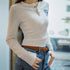Person wearing a beige long-sleeve shirt with a logo and blue jeans, standing indoors.